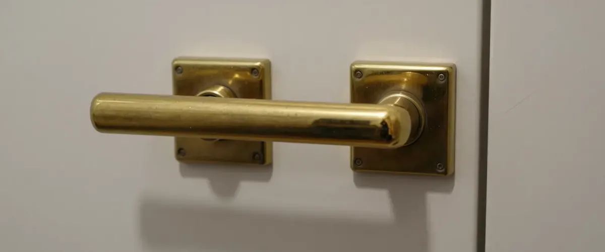 Goldfarbener Türgriff auf eckigen Messingplatten an einer glatten, hellen Tür, seitlich fällt weiches Licht ein